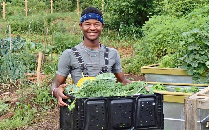 A person smiles at the camera while carrying a box filled with vegetables. A garden is in the background.